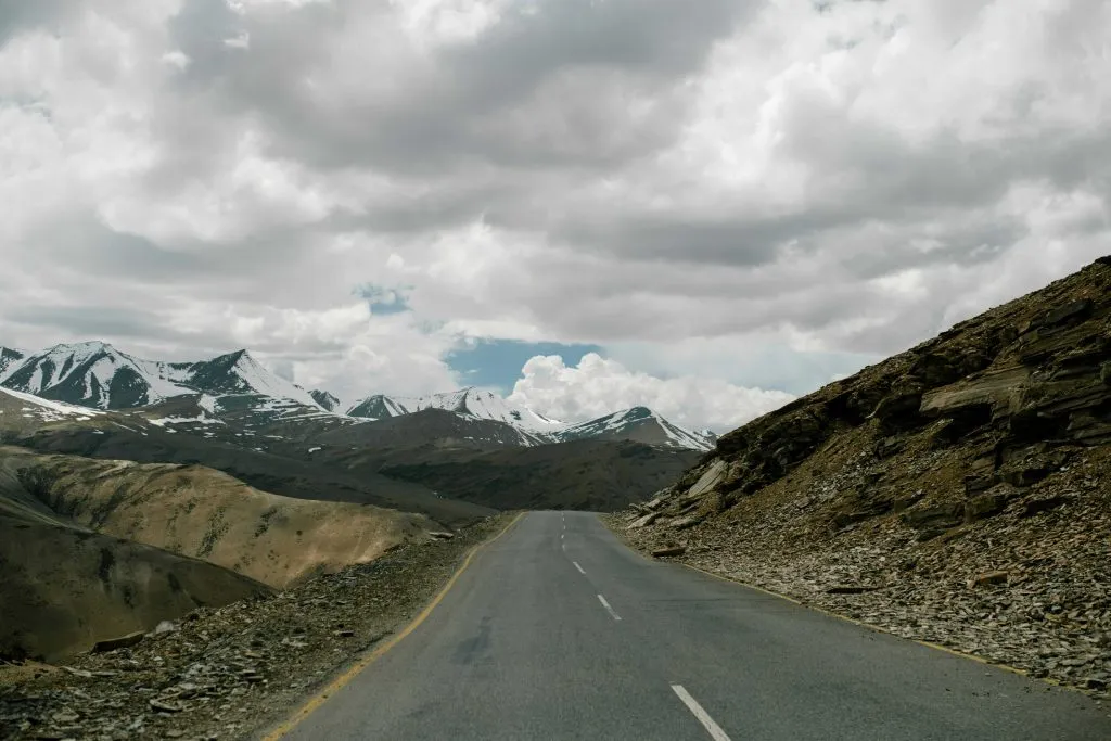 Beautiful road in Ladakh surrounded by rocky mountains and snow-capped Himalayan peaks under a dramatic cloudy sky, ideal for adventure travel and road trips